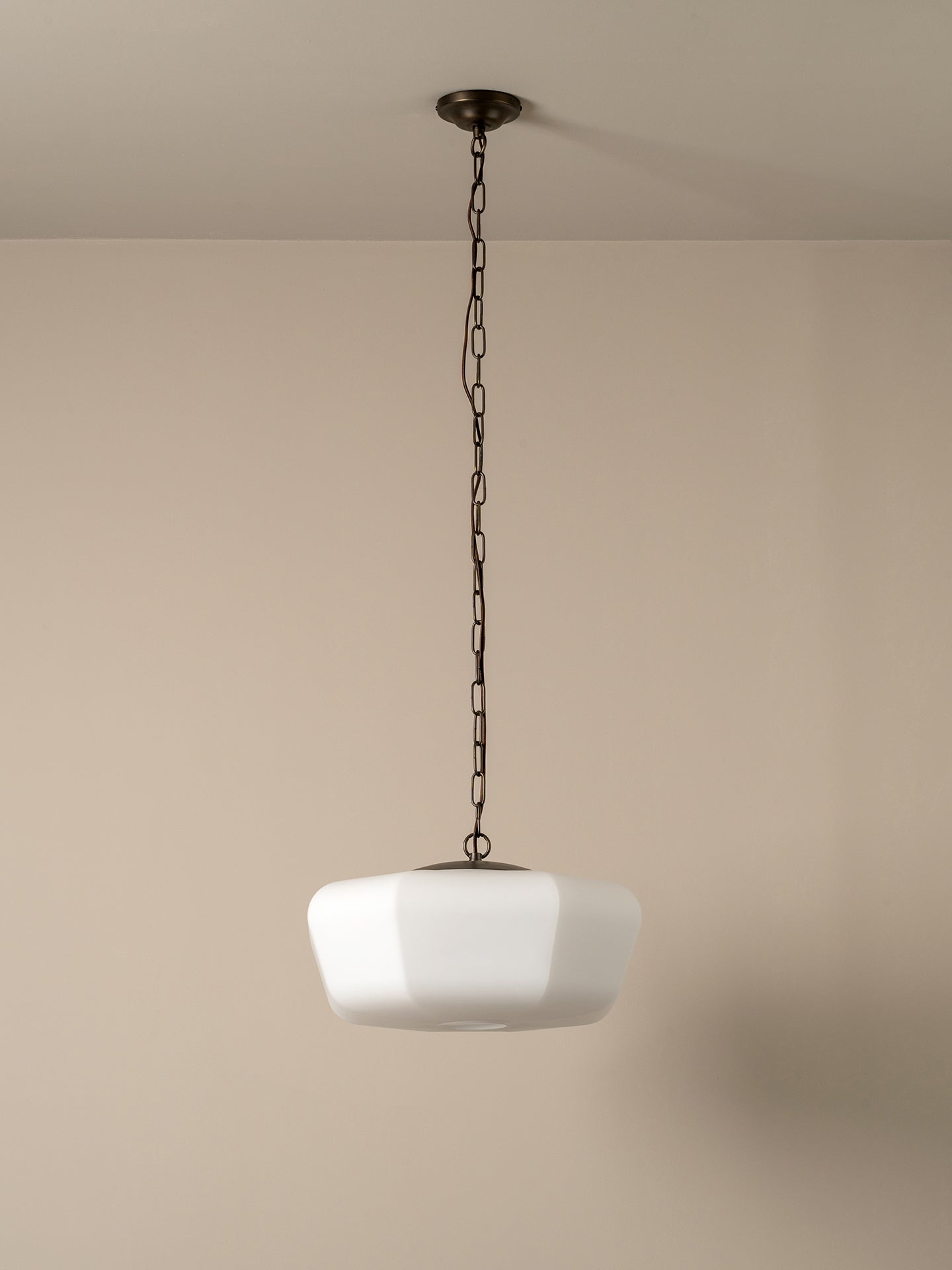 A modern pendant light with a white faceted glass shade suspended by a dark metal chain from a ceiling canopy, set against a minimalist beige wall.