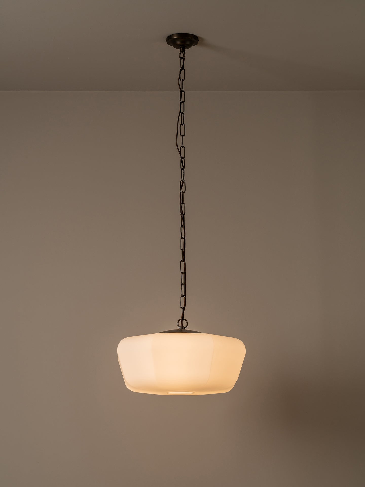 A modern pendant light with a warm white frosted glass shade suspended by a dark metal chain from a ceiling mount, set against a neutral beige wall.