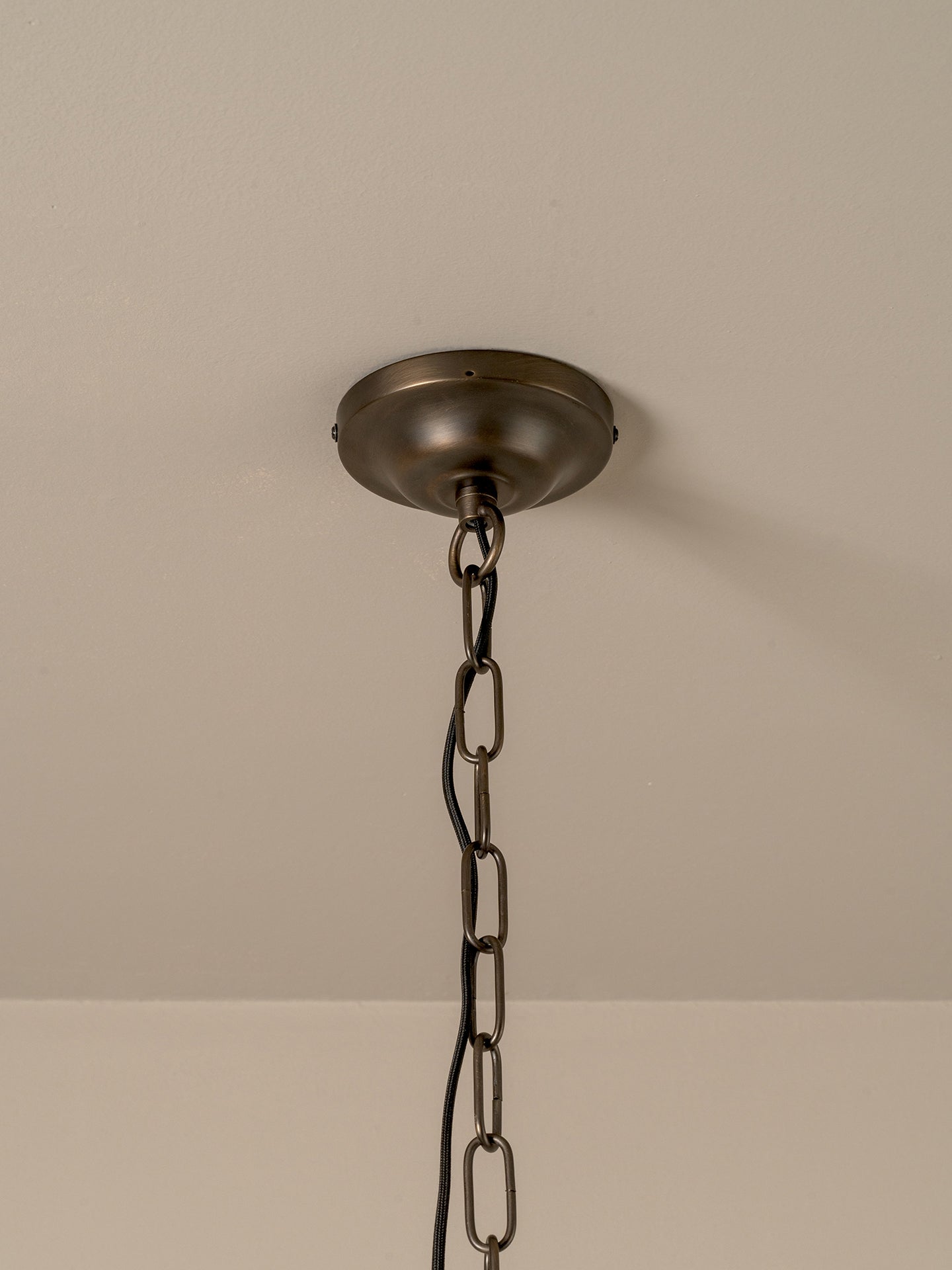 An industrial-style ceiling light fixture with a round, dark bronze ceiling canopy and a matching metal chain suspending the fixture against a plain beige wall and ceiling.