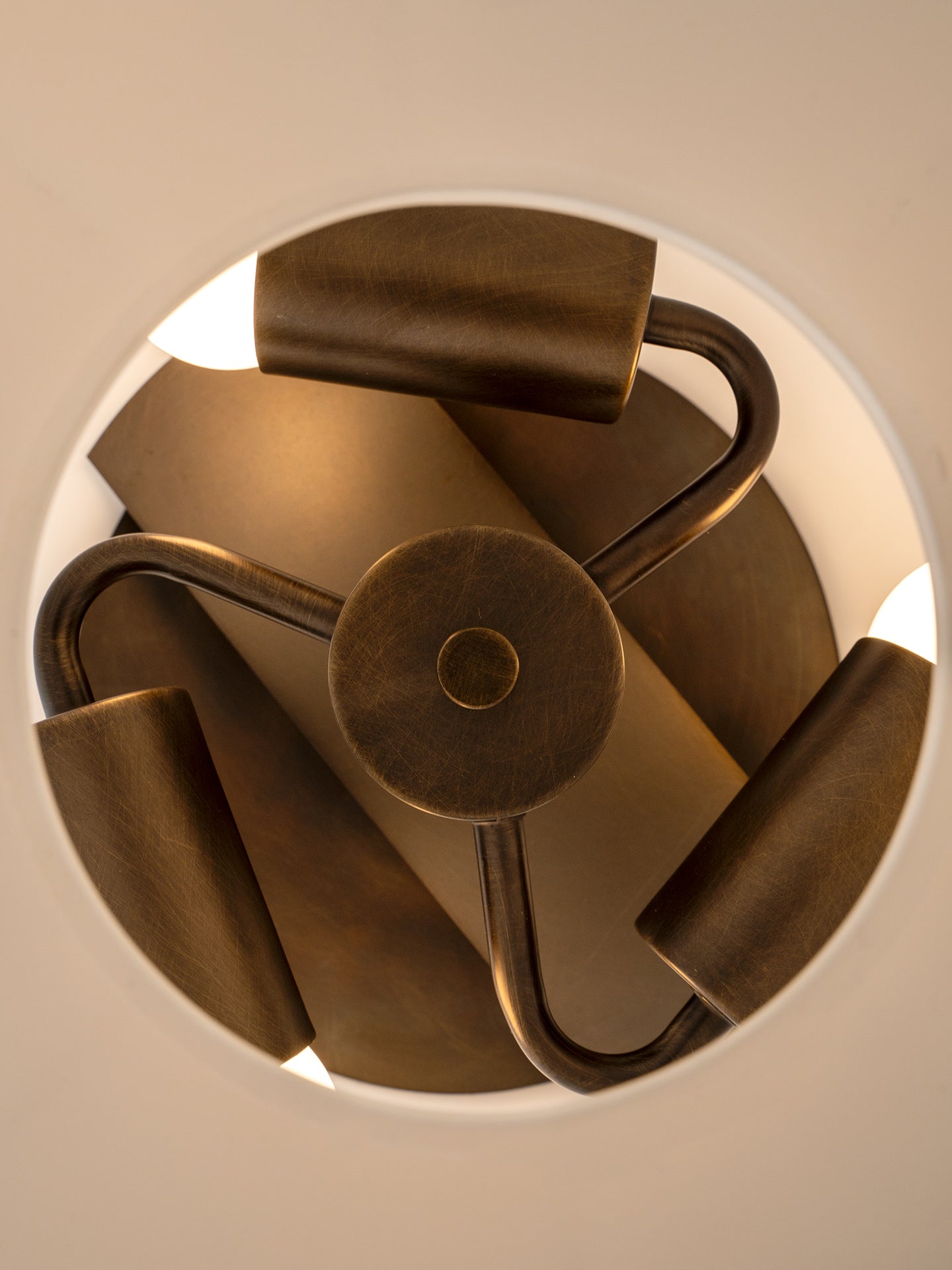 A close-up top-down view of a modern pendant light fixture with a circular frame, matte bronze metal arms, and cylindrical shades, glowing with warm light against a neutral background.