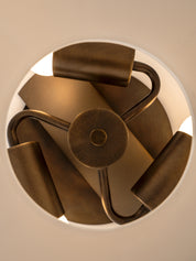 A close-up top-down view of a modern pendant light fixture with a circular frame, matte bronze metal arms, and cylindrical shades, glowing with warm light against a neutral background.