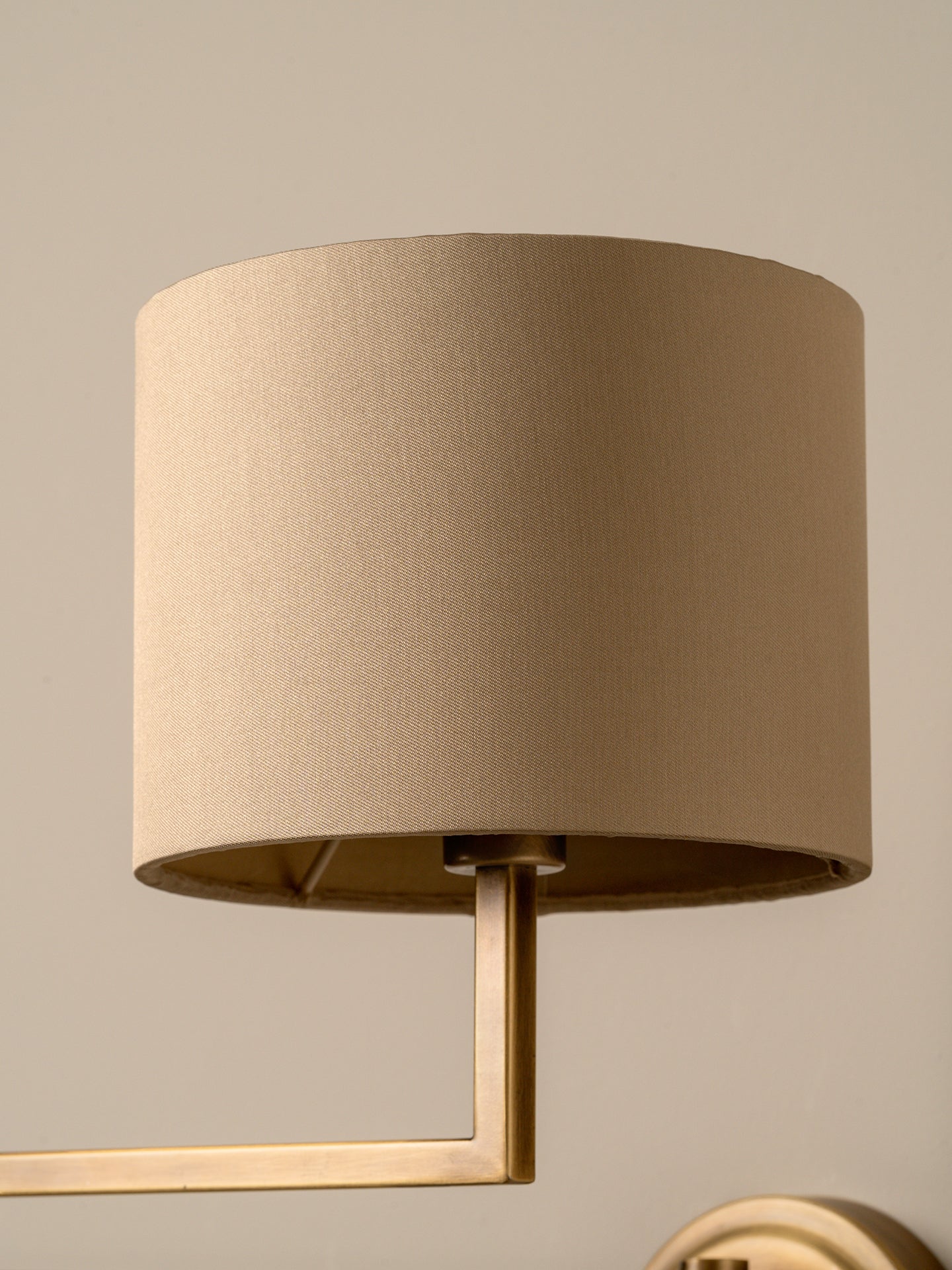 A close-up of a cylindrical fabric drum lampshade in a warm beige color, attached to a brass wall sconce with a matching beige background.