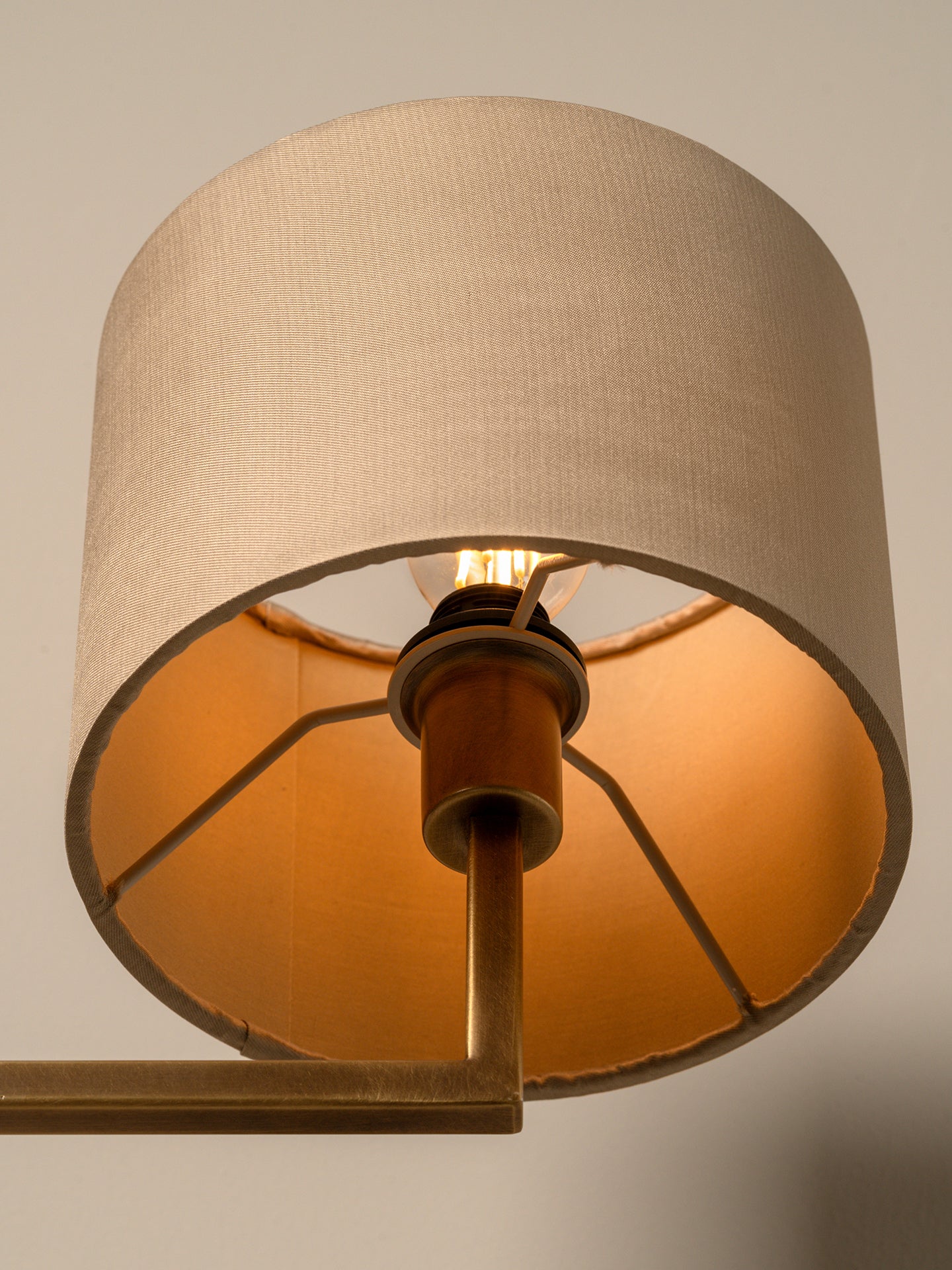 A close-up of a wall lamp with a large beige fabric drum shade, a warm orange-gold interior, and a brass metal fixture against a neutral light gray background.