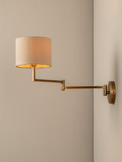 A modern wall-mounted swing-arm sconce with a cylindrical beige fabric shade and a warm brass-toned metal arm and wall plate, shown lit against a neutral beige wall.