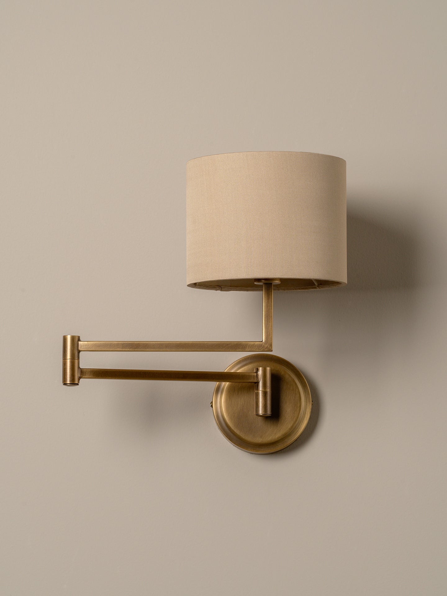 A modern wall-mounted sconce with a cylindrical beige fabric shade, adjustable brass arm, and round brass wall plate, shown against a plain warm beige wall.