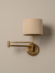 A modern wall-mounted sconce with a cylindrical beige fabric shade, adjustable brass arm, and round brass wall plate, shown against a plain warm beige wall.