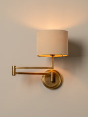 A modern wall-mounted swing-arm sconce with a cylindrical beige fabric shade and a circular brass base, shown lit against a plain beige wall.