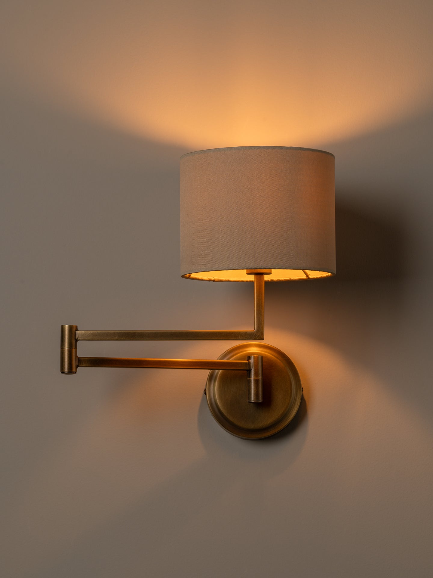 A wall-mounted adjustable sconce with a cylindrical beige fabric shade and a warm brass metal arm and base, shown lit against a neutral gray wall.