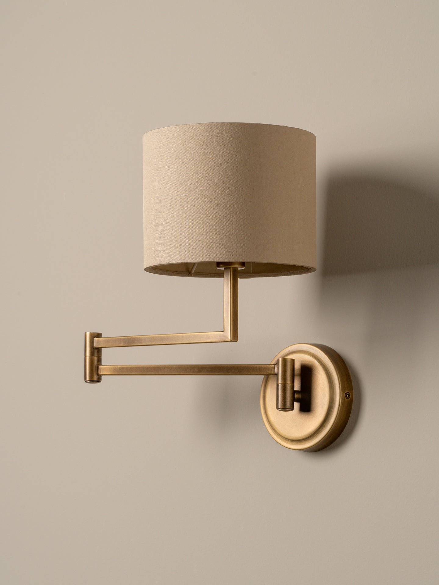 A wall-mounted adjustable sconce with a cylindrical beige fabric shade and a brushed brass metal arm and wall plate, shown against a plain beige wall.