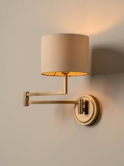 A modern wall-mounted sconce with a cylindrical beige fabric shade, a gold-finished adjustable metal arm, and a round wall plate, shown lit against a warm neutral wall.