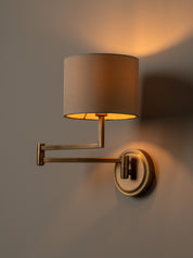 A modern wall-mounted swing-arm sconce with a cylindrical beige fabric shade and a circular wall plate, shown lit against a warm neutral wall.