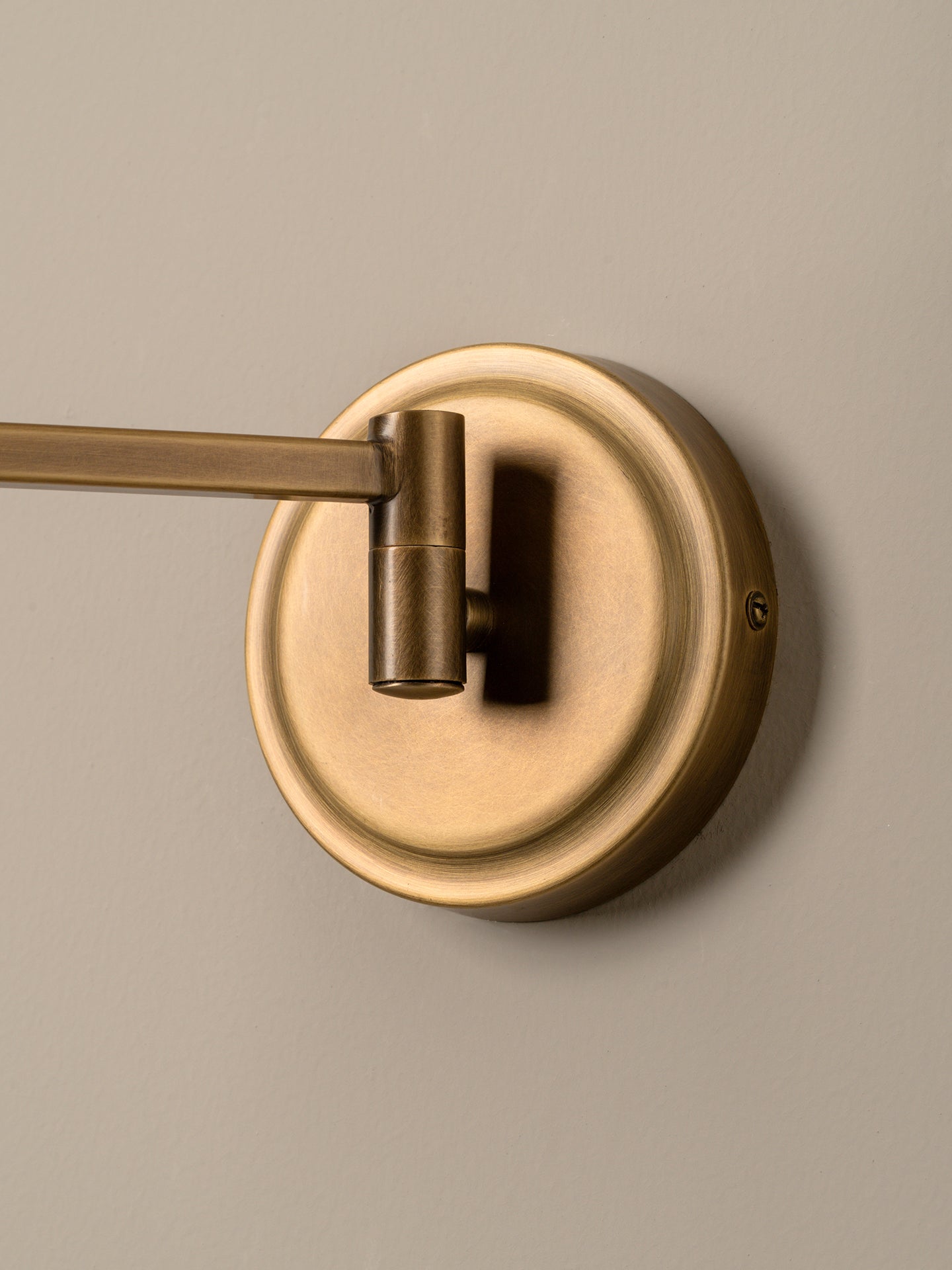 A close-up of a wall-mounted brass towel bar fixture with a round backplate and cylindrical swivel arm, shown against a neutral beige wall.