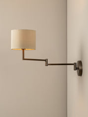 A modern wall-mounted swing-arm sconce with a cylindrical beige fabric shade and a dark metal arm, shown lit against a warm beige wall.