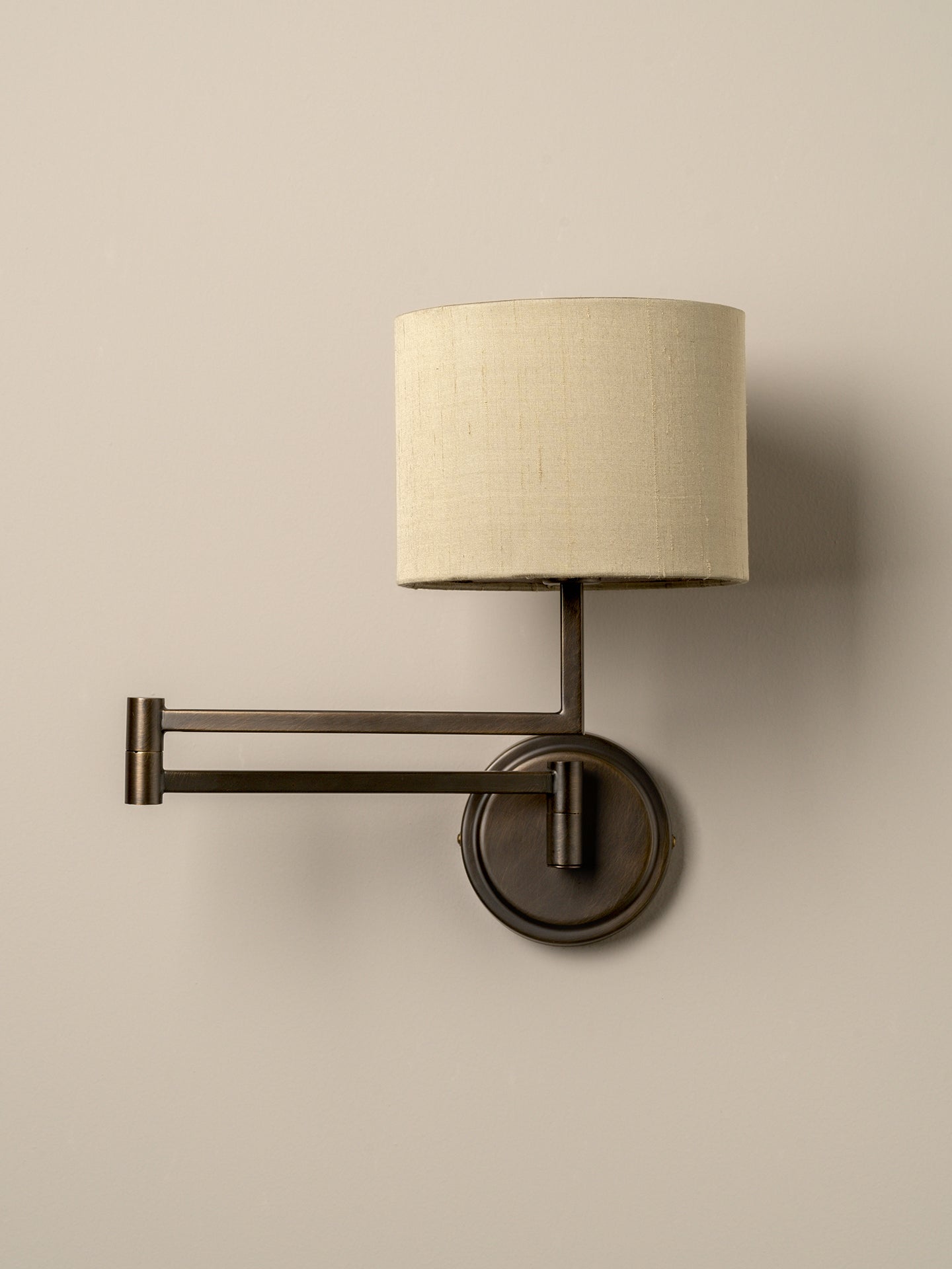 A wall-mounted swing-arm sconce with a cylindrical off-white linen shade and a dark bronze metal frame, shown against a plain beige wall.