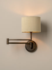 A wall-mounted swing-arm sconce with a cylindrical off-white fabric shade and a dark bronze metal frame, shown lit against a plain beige wall.