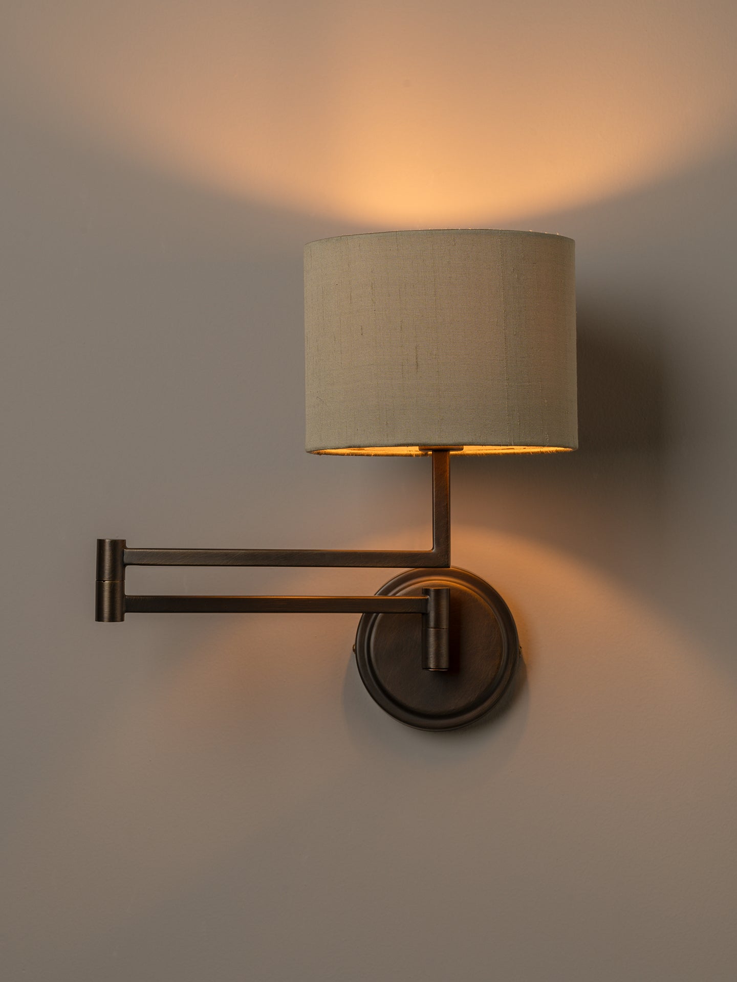 A wall-mounted adjustable sconce with a cylindrical beige fabric shade and a dark metal arm and base, shown lit against a plain wall.