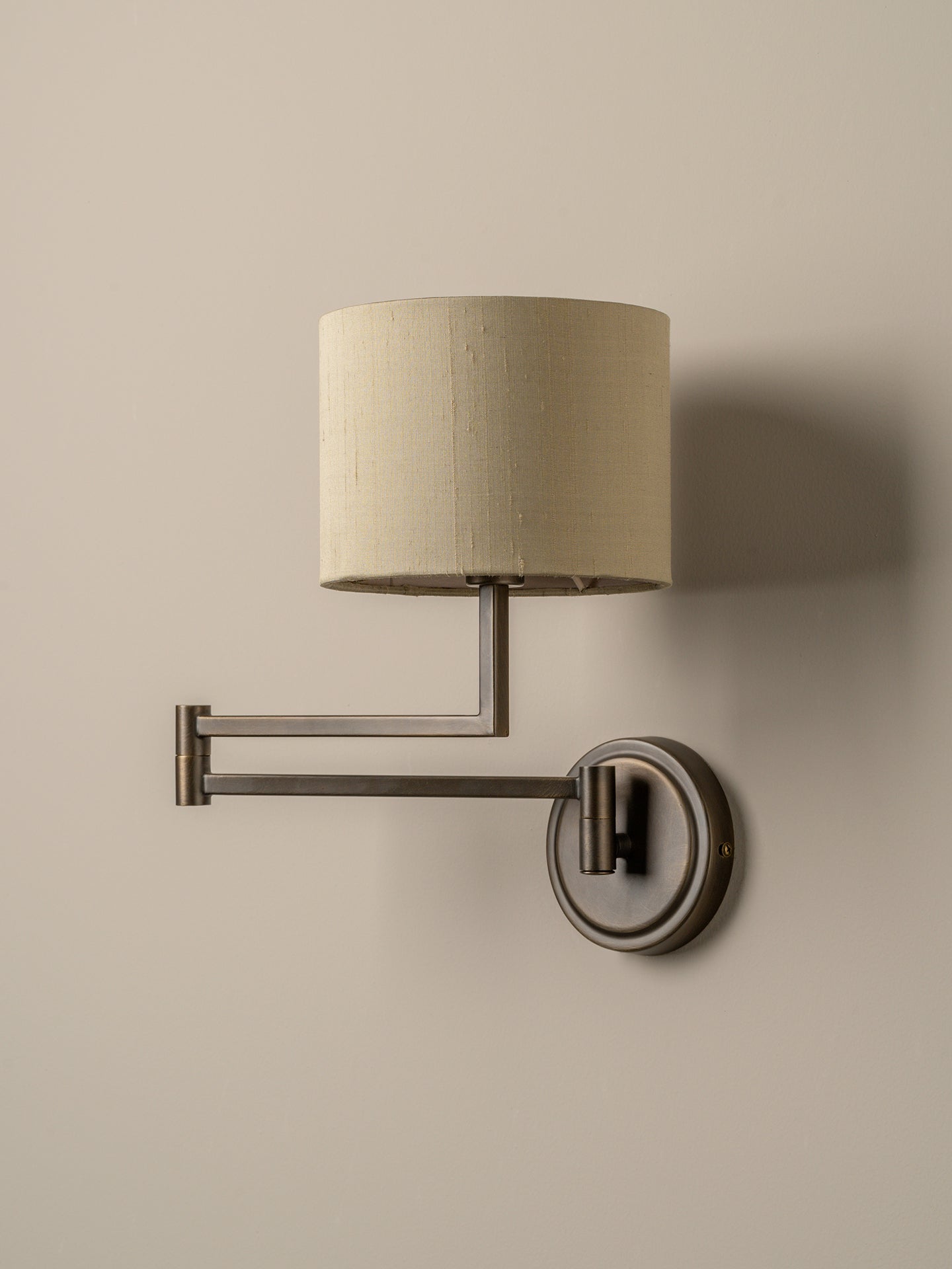 A modern wall-mounted swing-arm sconce with a cylindrical beige fabric shade and a dark brushed metal frame, mounted on a plain beige wall.