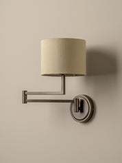 A modern wall-mounted swing-arm sconce with a cylindrical beige fabric shade and a dark brushed metal frame, mounted on a plain beige wall.