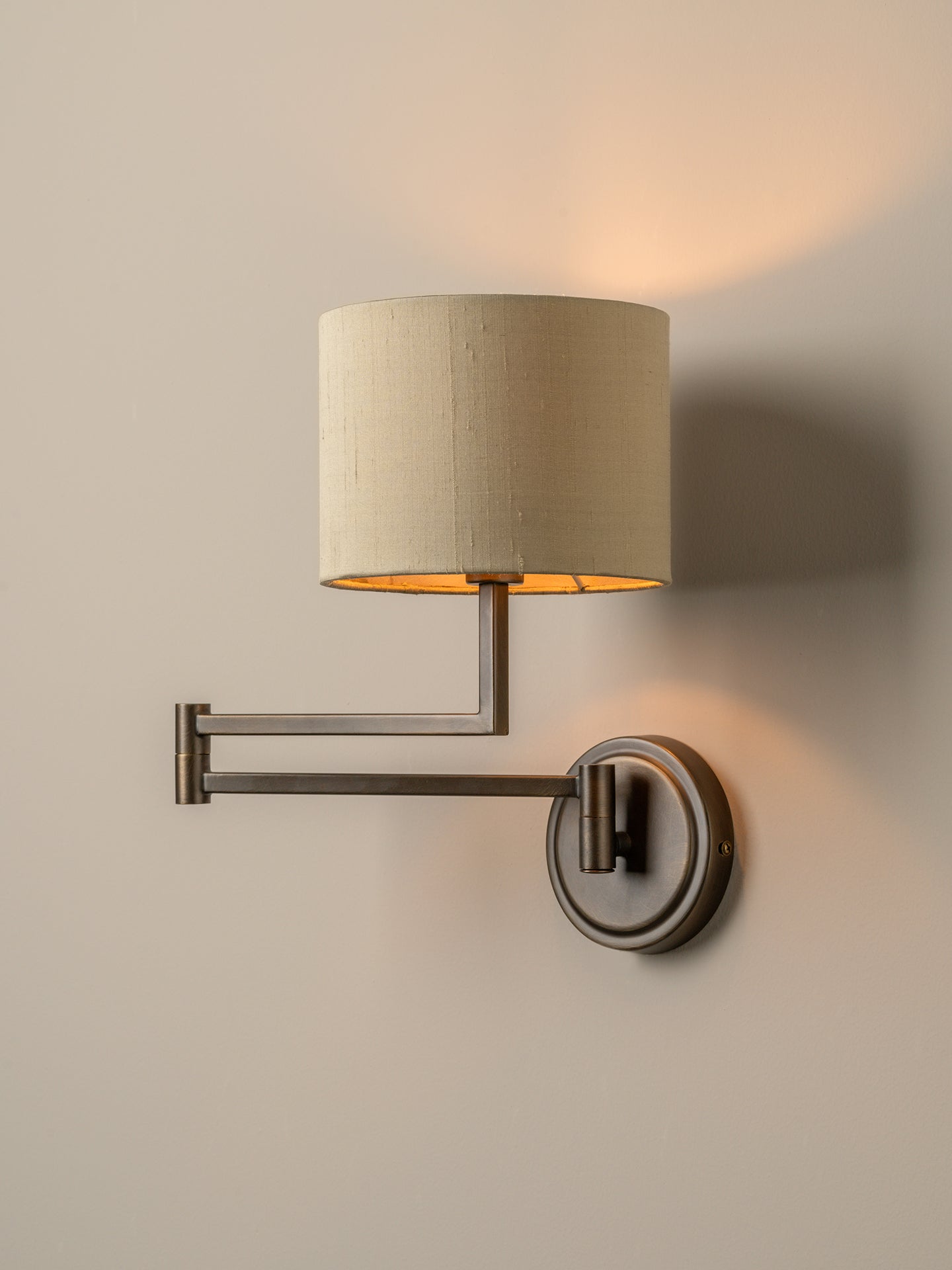 A modern wall-mounted sconce with a cylindrical beige fabric shade, adjustable metal arm, and circular wall mount, shown lit against a warm neutral wall.