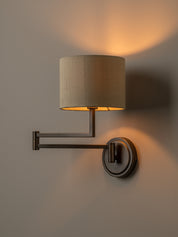 A modern wall-mounted sconce with a cylindrical beige fabric shade and a dark metal adjustable arm, shown lit against a neutral wall.