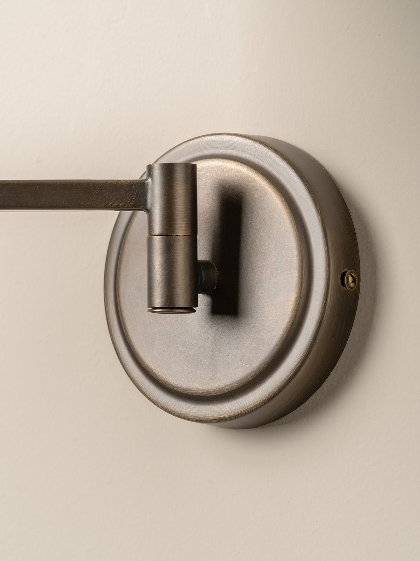 A close-up of a wall-mounted light fixture with a circular backplate and a horizontal arm in a brushed metal finish, mounted on a plain beige wall.