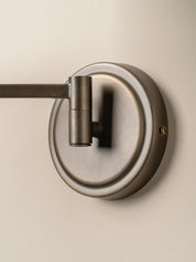 A close-up of a wall-mounted light fixture with a circular backplate and a horizontal arm in a brushed metal finish, mounted on a plain beige wall.