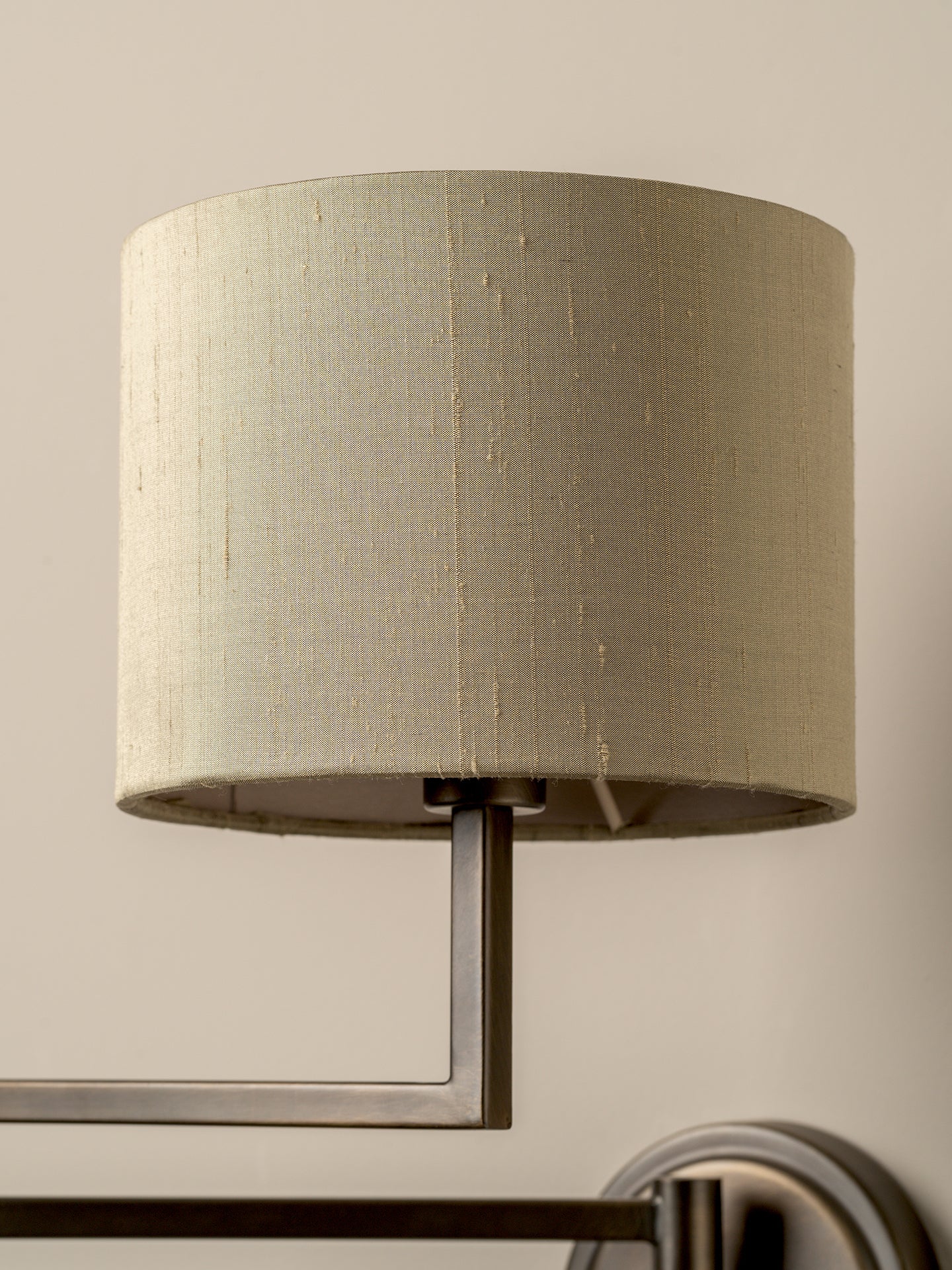 A close-up of a cylindrical drum lampshade in a textured beige fabric, mounted on a brushed metal wall sconce against a warm beige wall.