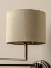 A close-up of a cylindrical drum lampshade in a textured beige fabric, mounted on a brushed metal wall sconce against a warm beige wall.