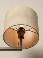 A close-up of a cylindrical drum lampshade in a textured off-white linen fabric, with a warm amber light glowing from within. The shade is mounted on a dark metal wall sconce with a visible light bulb inside.