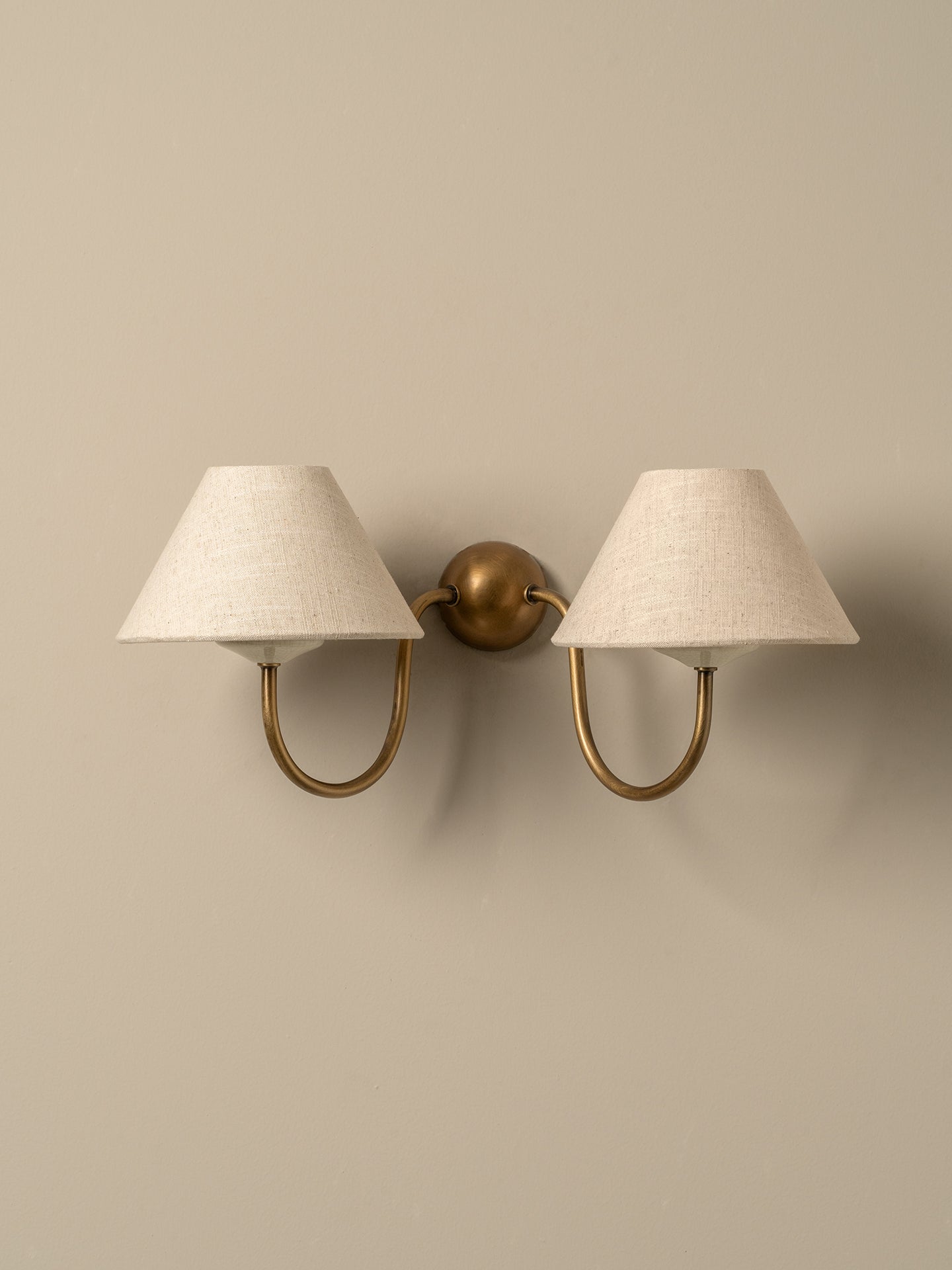 A two-light wall sconce with tapered off-white fabric shades and curved brass arms, mounted on a plain beige wall.