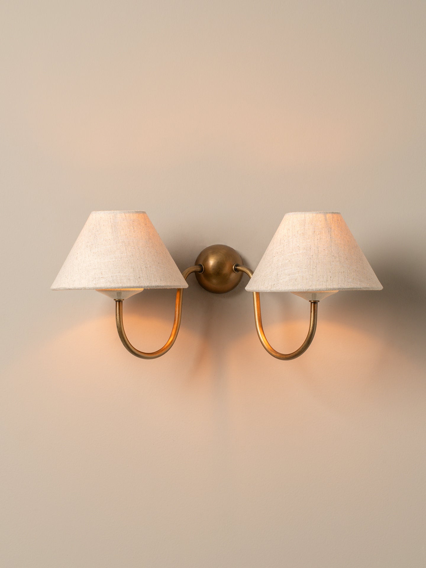 A two-light wall sconce with textured off-white fabric shades and curved brass arms, mounted on a warm beige wall and casting a soft glow.