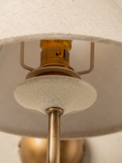 Close-up detail of a table lamp showing a cream fabric drum shade, a white crackled glass diffuser, and an antique brass metal socket and stem.