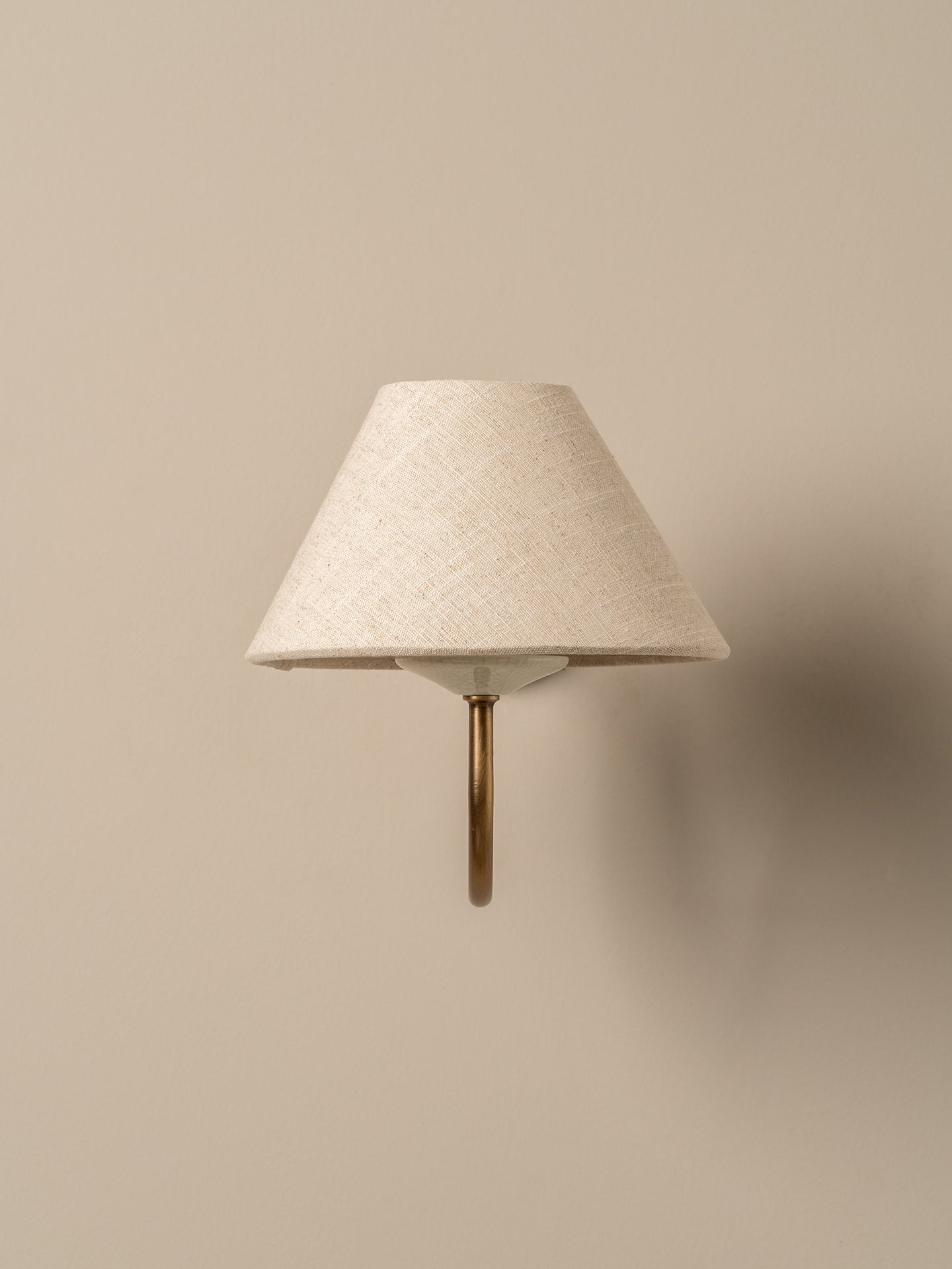A modern wall-mounted sconce with a textured cream fabric cone shade and a curved brass metal stem, shown against a solid beige wall.