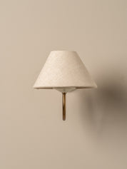 A modern wall-mounted sconce with a textured cream fabric cone shade and a curved brass metal stem, shown against a solid beige wall.