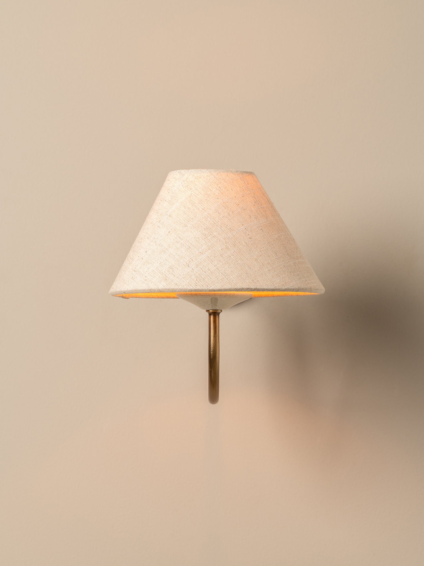 A modern wall-mounted sconce with a tapered off-white linen shade and a slim brass metal stem, casting a warm light against a neutral beige wall.
