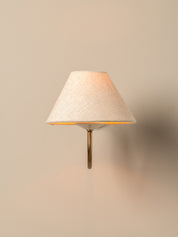 A modern wall-mounted sconce with a tapered off-white linen shade and a slim brass metal stem, casting a warm light against a neutral beige wall.