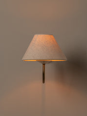 A modern wall-mounted sconce with a textured beige fabric drum shade and a slim brass metal stem, illuminated with a warm orange glow against a neutral beige wall.