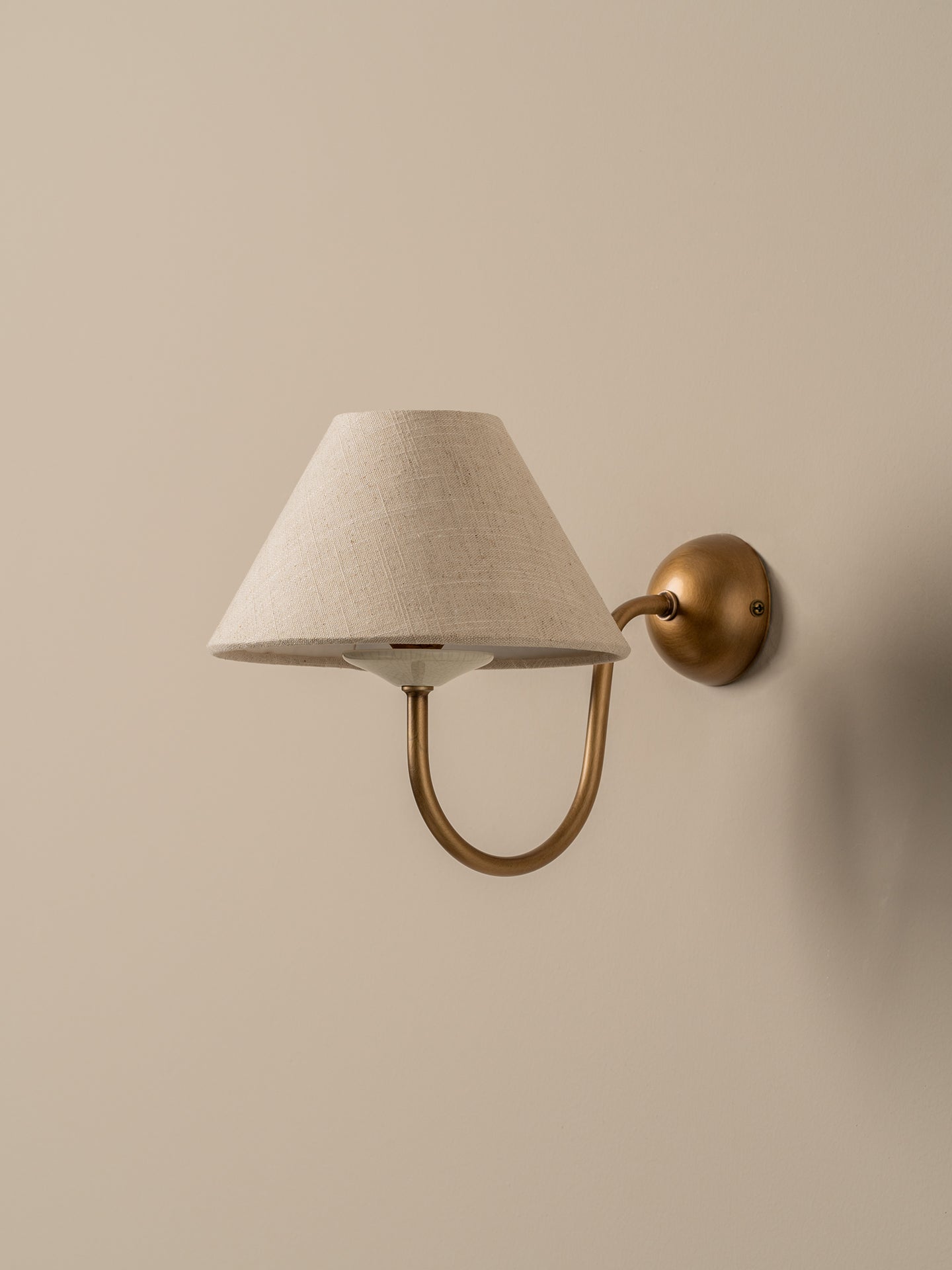 A wall-mounted sconce with a tapered off-white fabric shade and a curved brass arm, mounted on a beige wall.