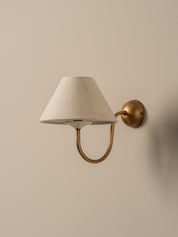 A wall-mounted sconce with a tapered off-white fabric shade and a curved brass arm, mounted on a beige wall.