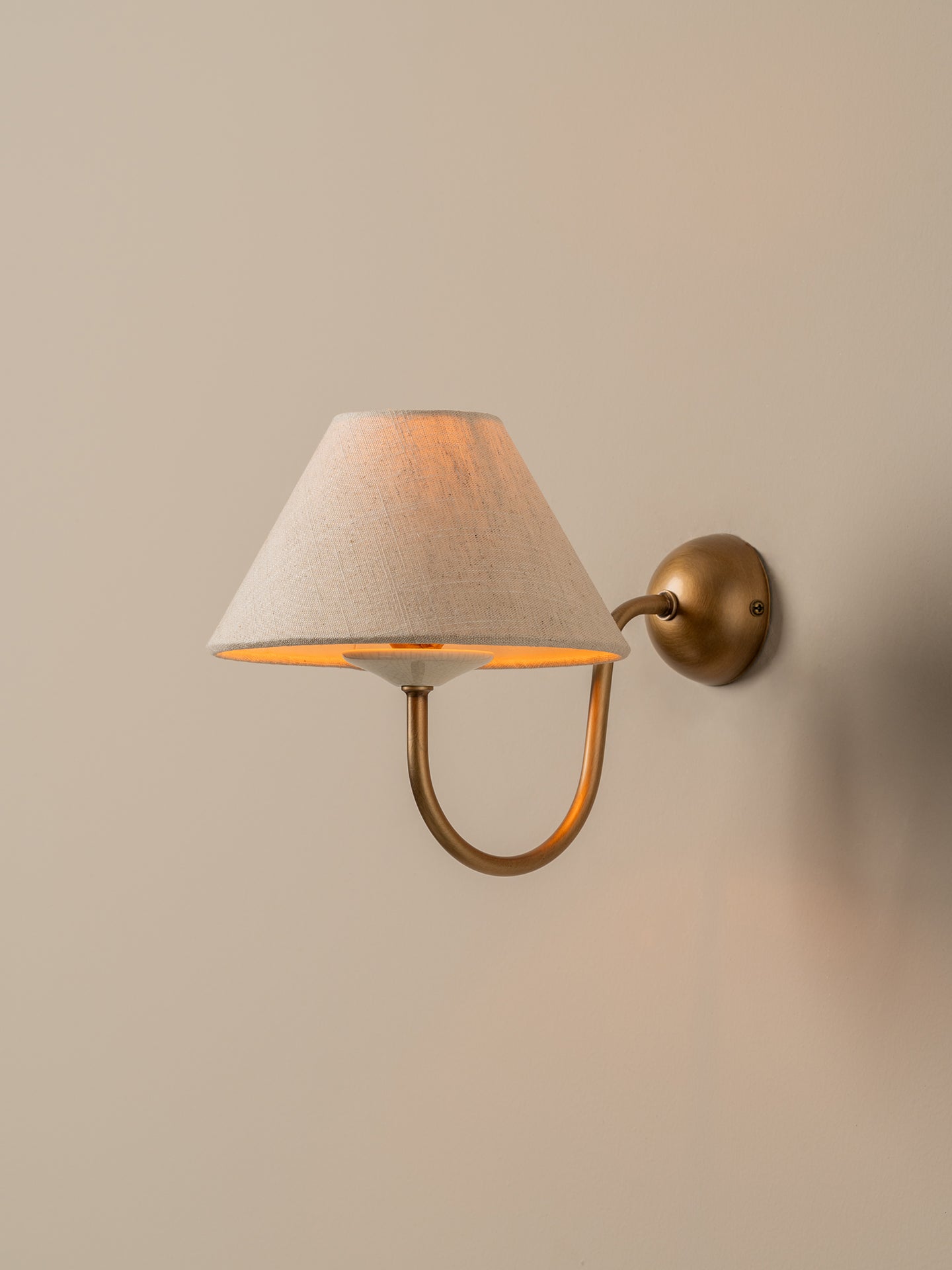 A wall-mounted sconce with a tapered off-white fabric shade and a curved brass arm, mounted on a beige wall.
