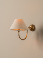 A wall-mounted sconce with a tapered off-white fabric shade and a curved brass arm, mounted on a beige wall.