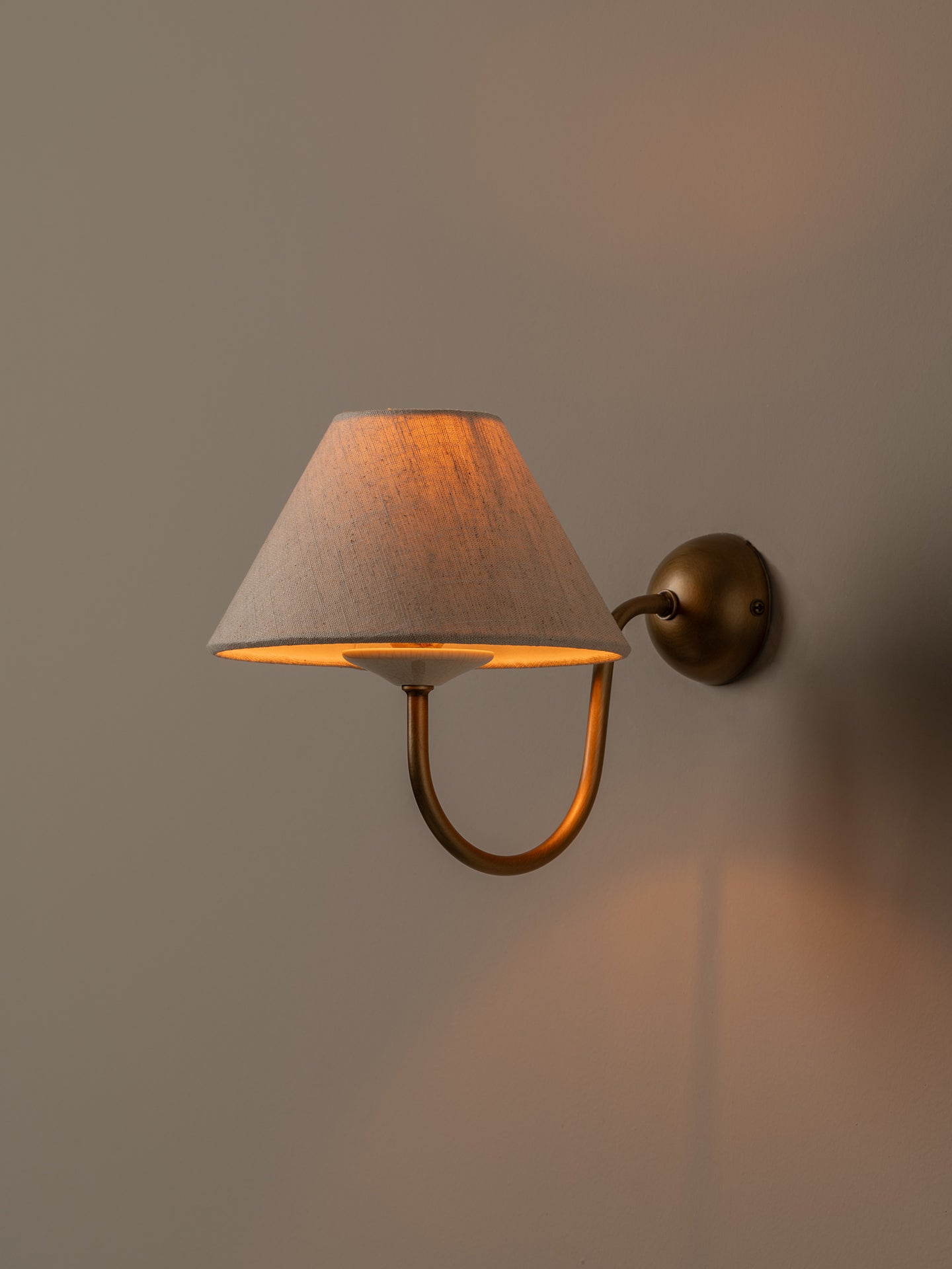 A wall-mounted sconce with a tapered beige fabric shade and a curved brass-toned metal arm, shown lit against a warm neutral wall.