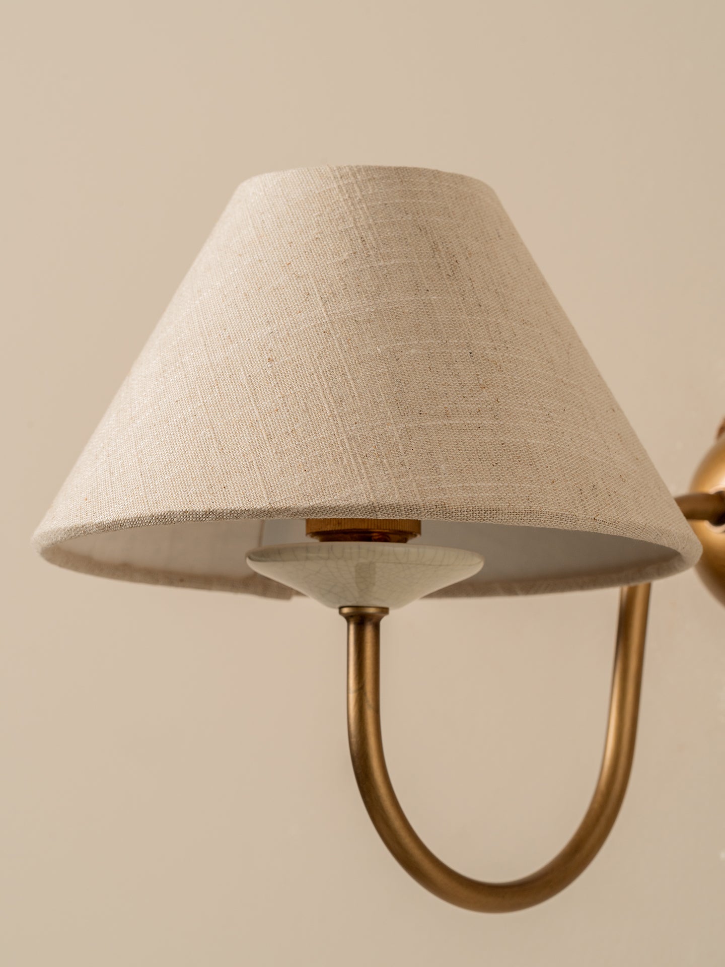 A close-up of a wall-mounted lamp with a tapered beige linen shade and a curved brass metal arm against a warm beige background.