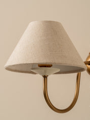 A close-up of a wall-mounted lamp with a tapered beige linen shade and a curved brass metal arm against a warm beige background.