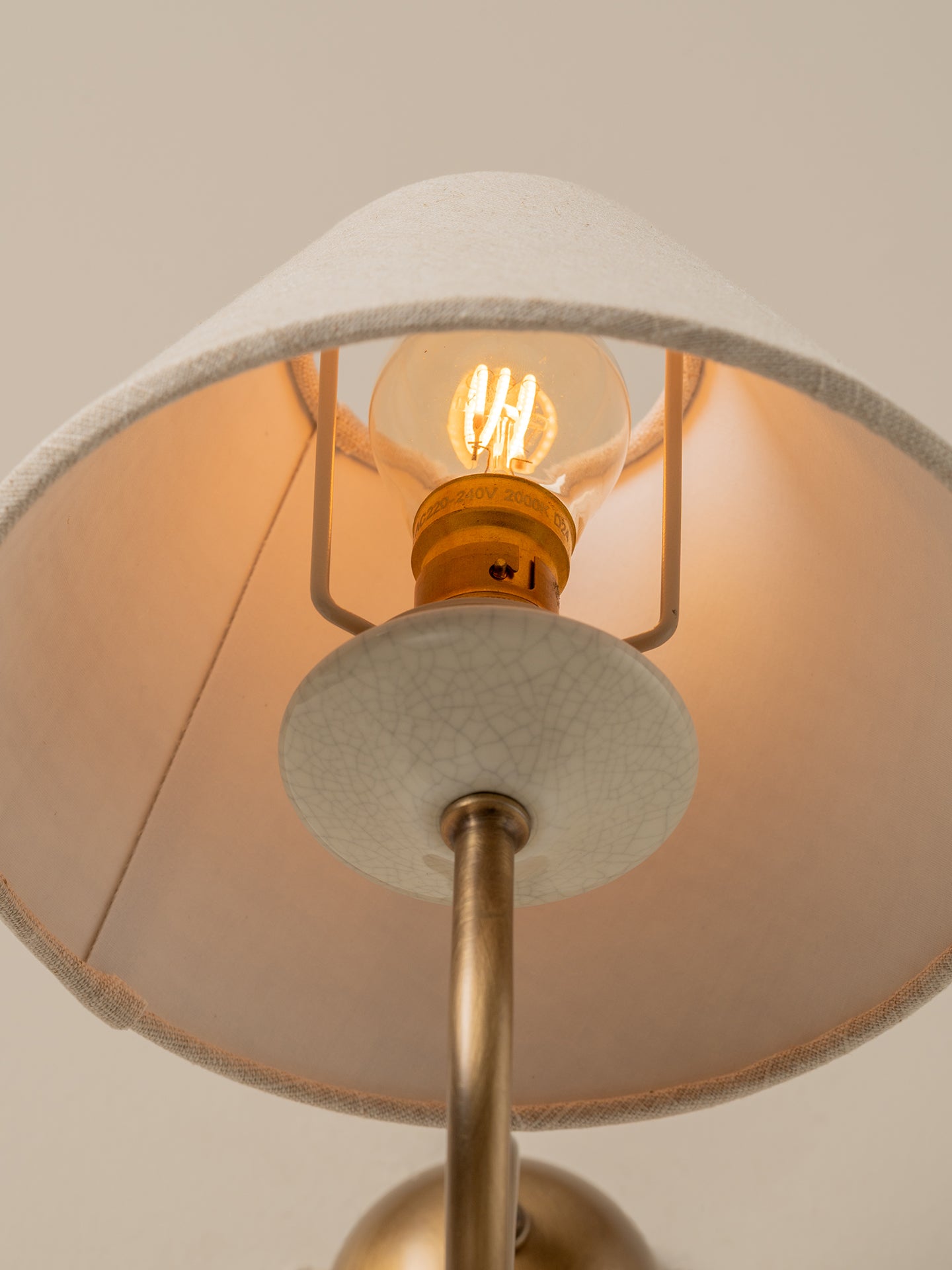 Close-up of a table lamp with a warm glowing Edison bulb, a cream fabric shade, and a brass metal stem against a neutral beige background.