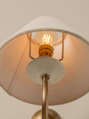 Close-up of a table lamp with a warm glowing Edison bulb, a cream fabric shade, and a brass metal stem against a neutral beige background.