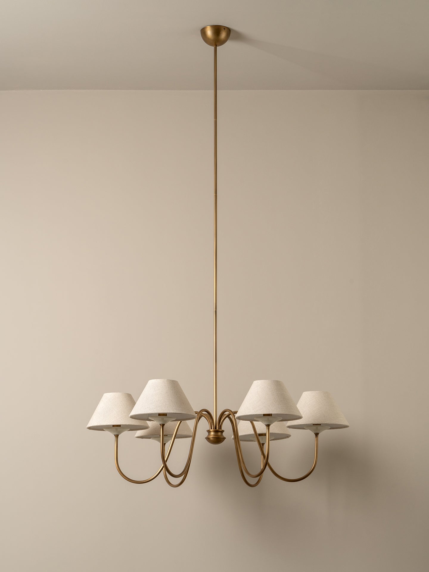 A modern six-light chandelier with a brushed brass or gold-finished metal frame and tapered off-white fabric shades, suspended from a matching ceiling canopy against a warm beige wall.