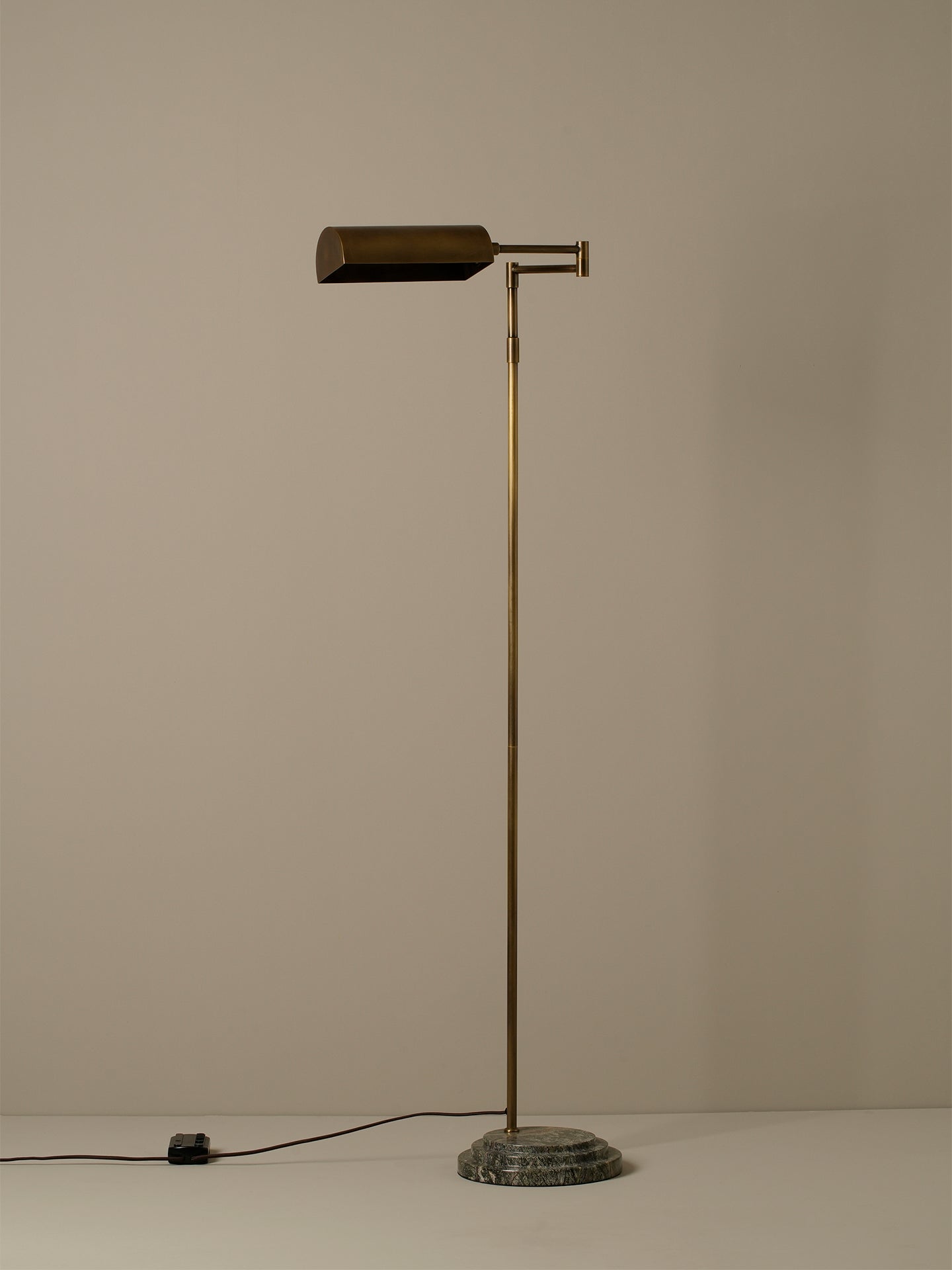 A vintage-style floor lamp with a cylindrical brass shade, a slender brass pole, and a round dark stone base, photographed against a plain beige wall.