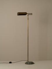 A vintage-style floor lamp with a cylindrical brass shade, a slender brass pole, and a round dark stone base, photographed against a plain beige wall.