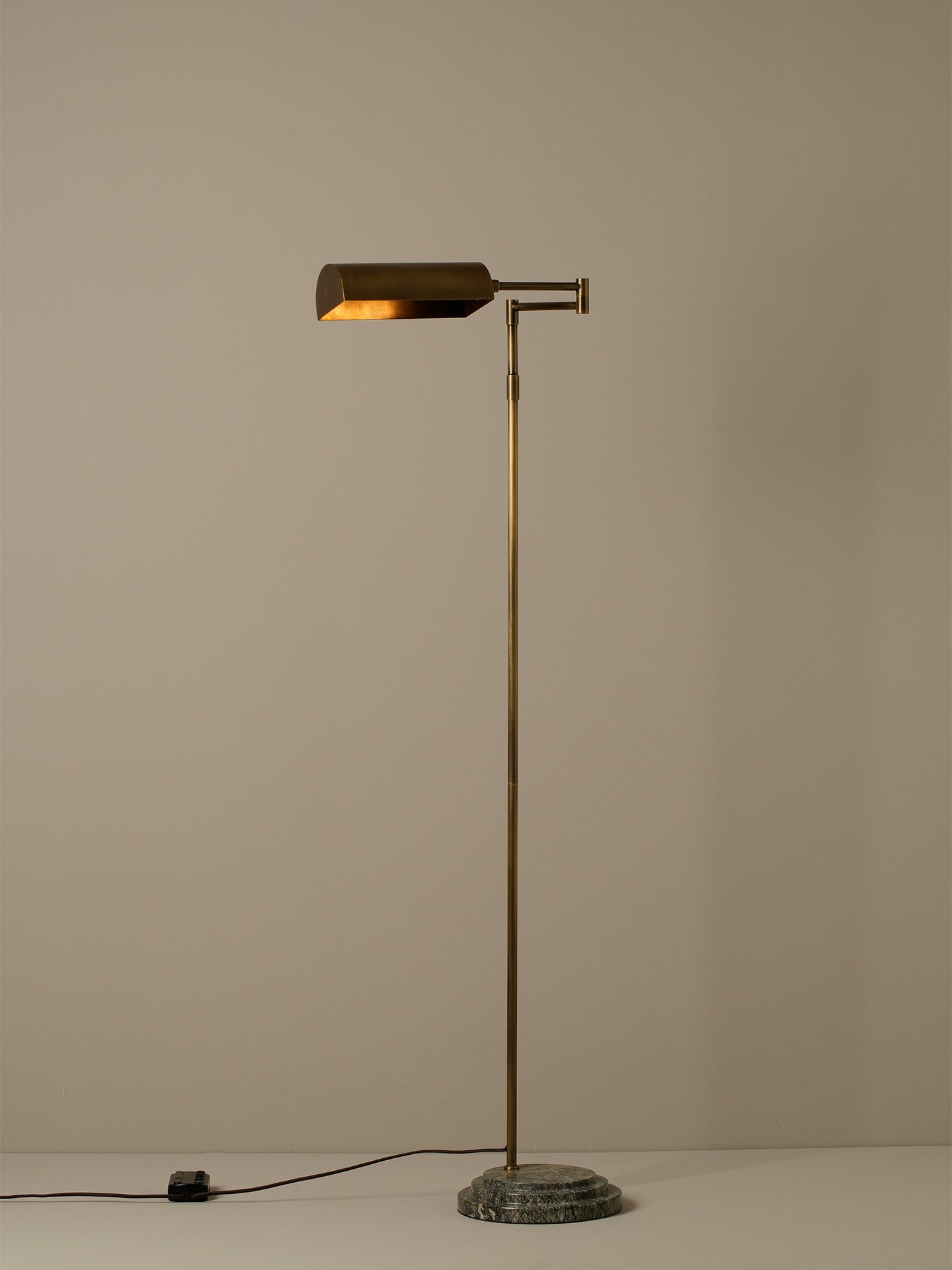 A modern floor lamp with a brass-finished pole and adjustable rectangular shade, mounted on a round dark stone base, photographed against a plain warm beige wall.